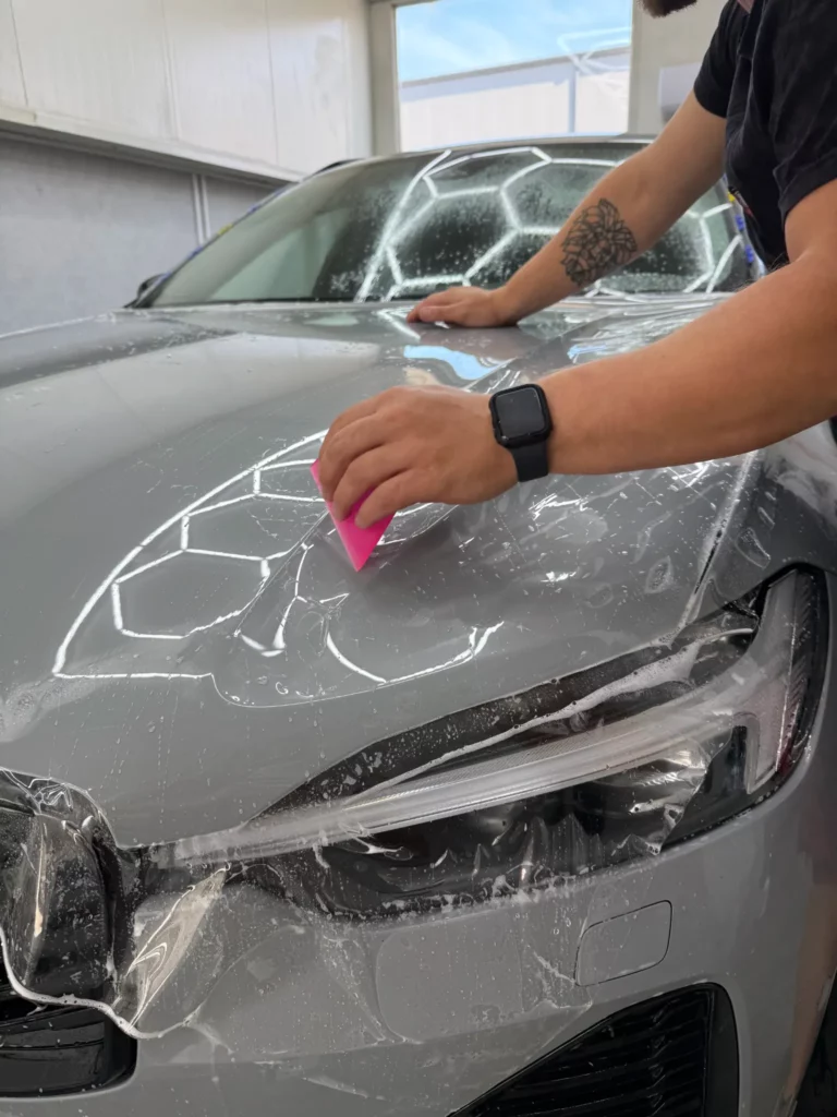 Zbliżenie dłoni z raklą podczas aplikacji PPF na masce Volvo XC60 nad reflektorem